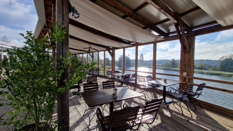 Terrasse, © Die Käsemacherwelt Überdachte Terrasse mit Holztischen und Stühlen am Wasser.