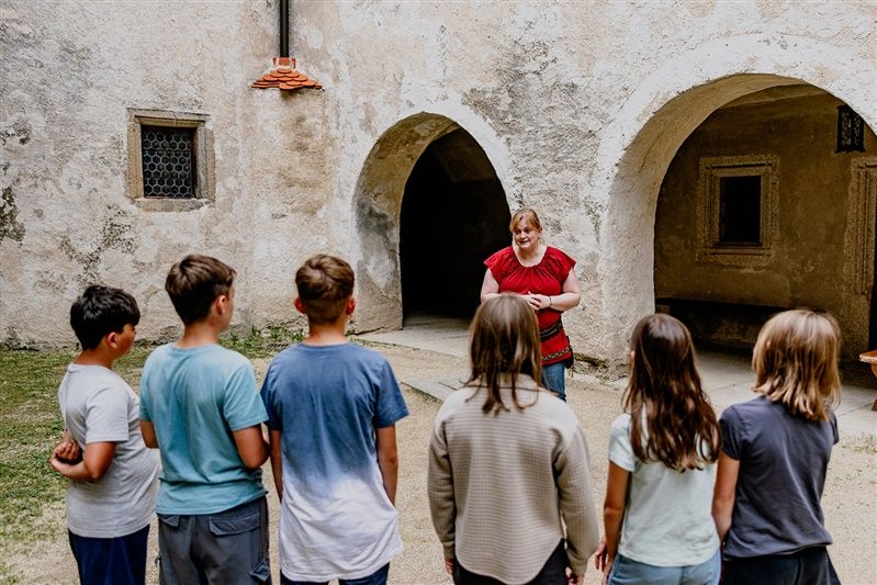 Eine Frau in rotem Oberteil spricht zu einer Gruppe von Kindern vor einer alten Steinmauer mit Torbögen.