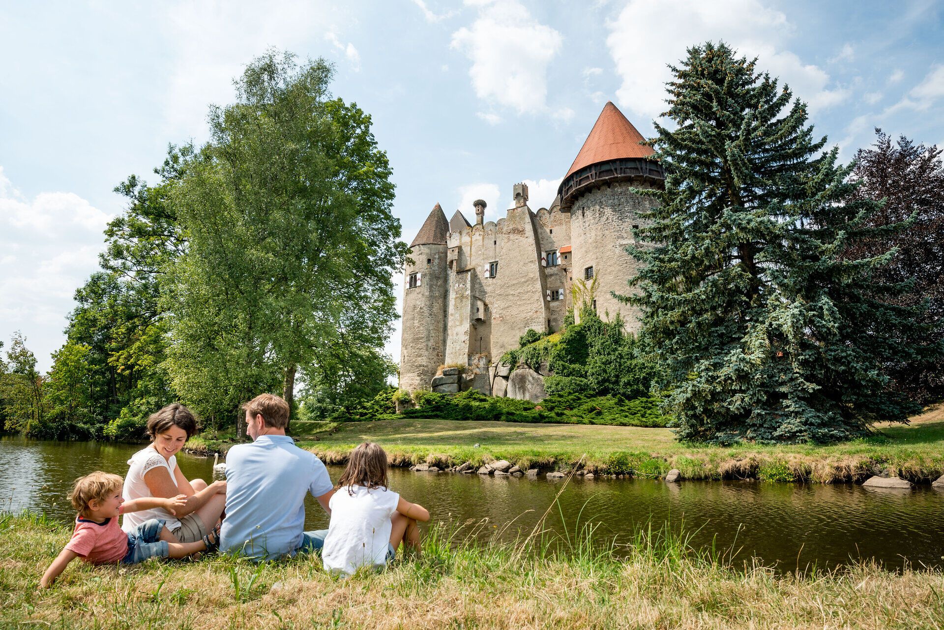 Die malerische Burg Heidenreichstein thront majestätisch über dem ruhigen Gewässer, während eine Familie entspannt im Gras sitzt und die Schönheit der Natur genießt. Umgeben von üppigem Grün und hohen Bäumen, strahlt der Ort eine friedliche Atmosphäre aus, die zum Verweilen einlädt.