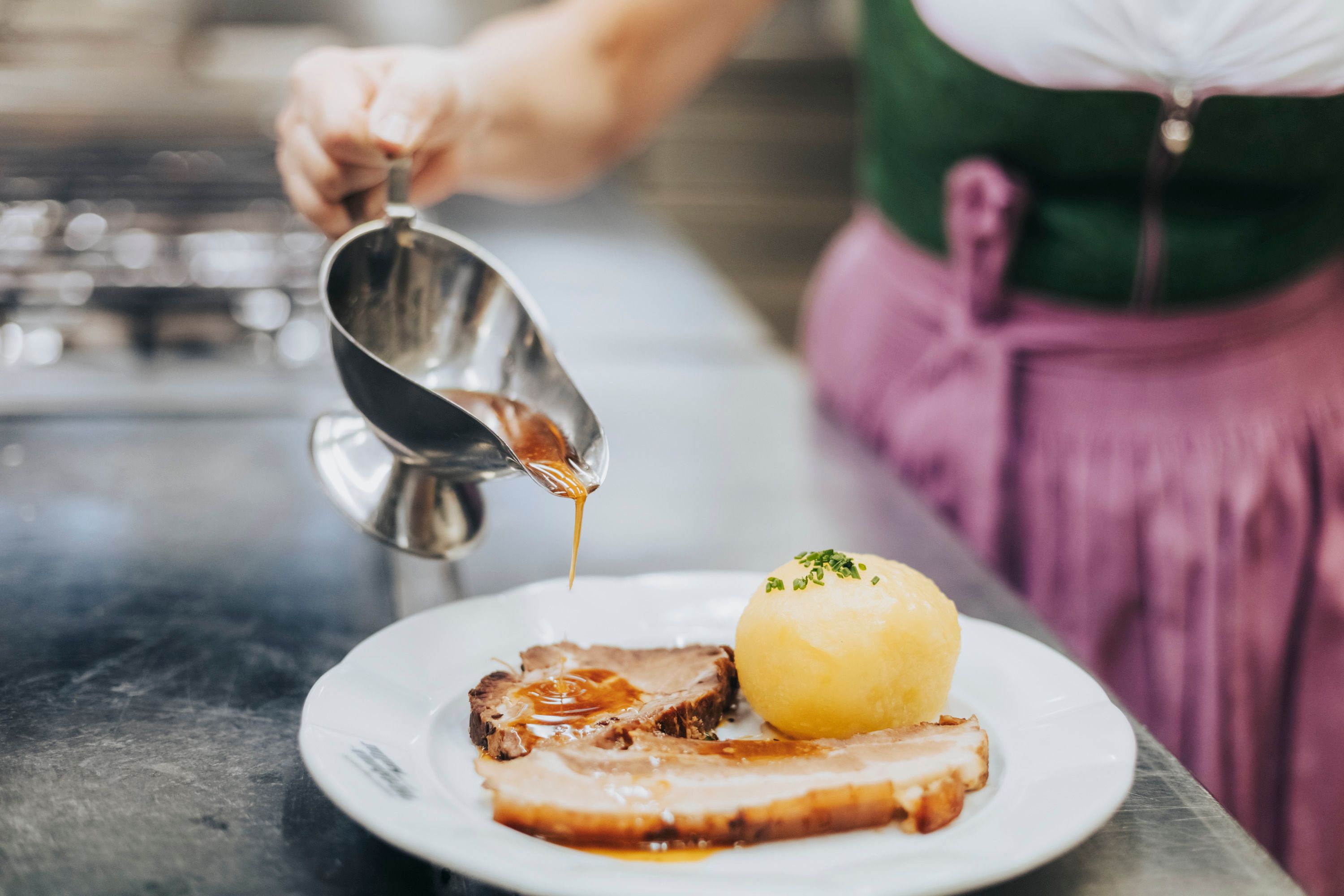 Eine Person gießt Soße über einen Teller mit Braten und Knödel.