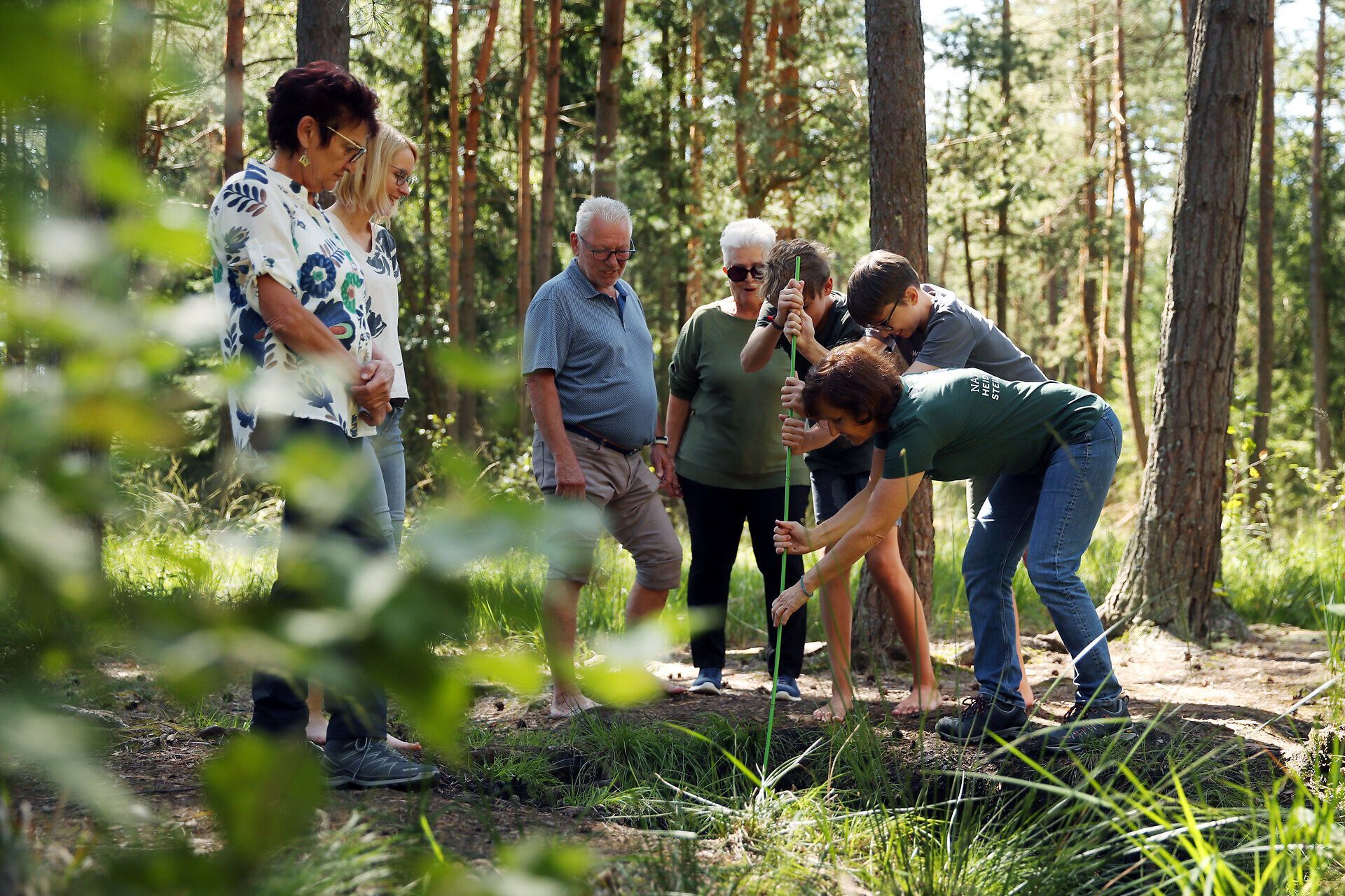Eine Gruppe, begleitet von einer Naturvermittlerin steht rund um ein Moorbecken. Zwei Jungen versuchen gemeinsam mit einer Naturvermittlerin die Tiefe des Moors zu messen.