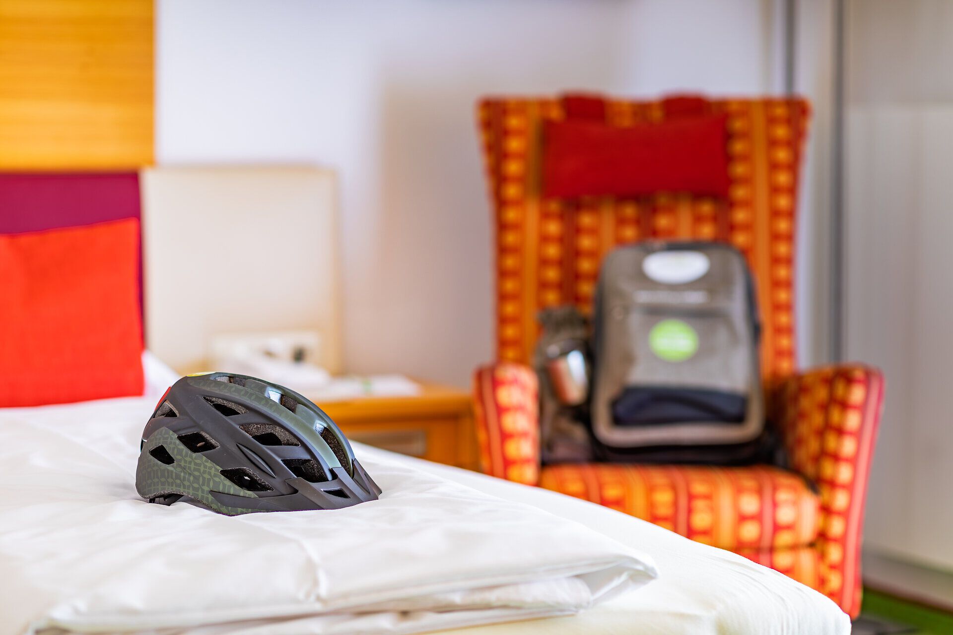Ein Fahrradhelm liegt auf dem Bett in einem Zimmer im Vitalzeit Hotel Weber. Dahinter steht ein rot-oranger gemütlicher Sessel, auf dem ein Rucksack abgestellt wurde.