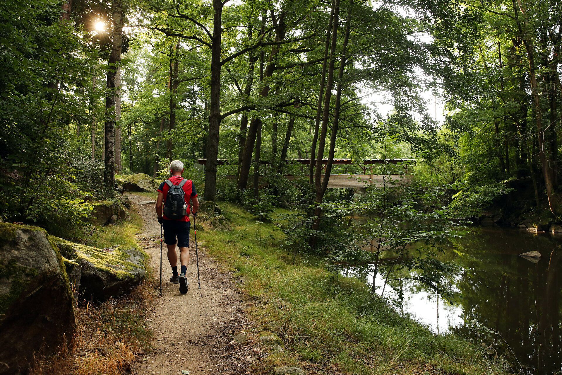 Ein Mann wandert einen Fluss entlang. Im Hintergrund ist eine große hölzerne Brücke zu sehen. 