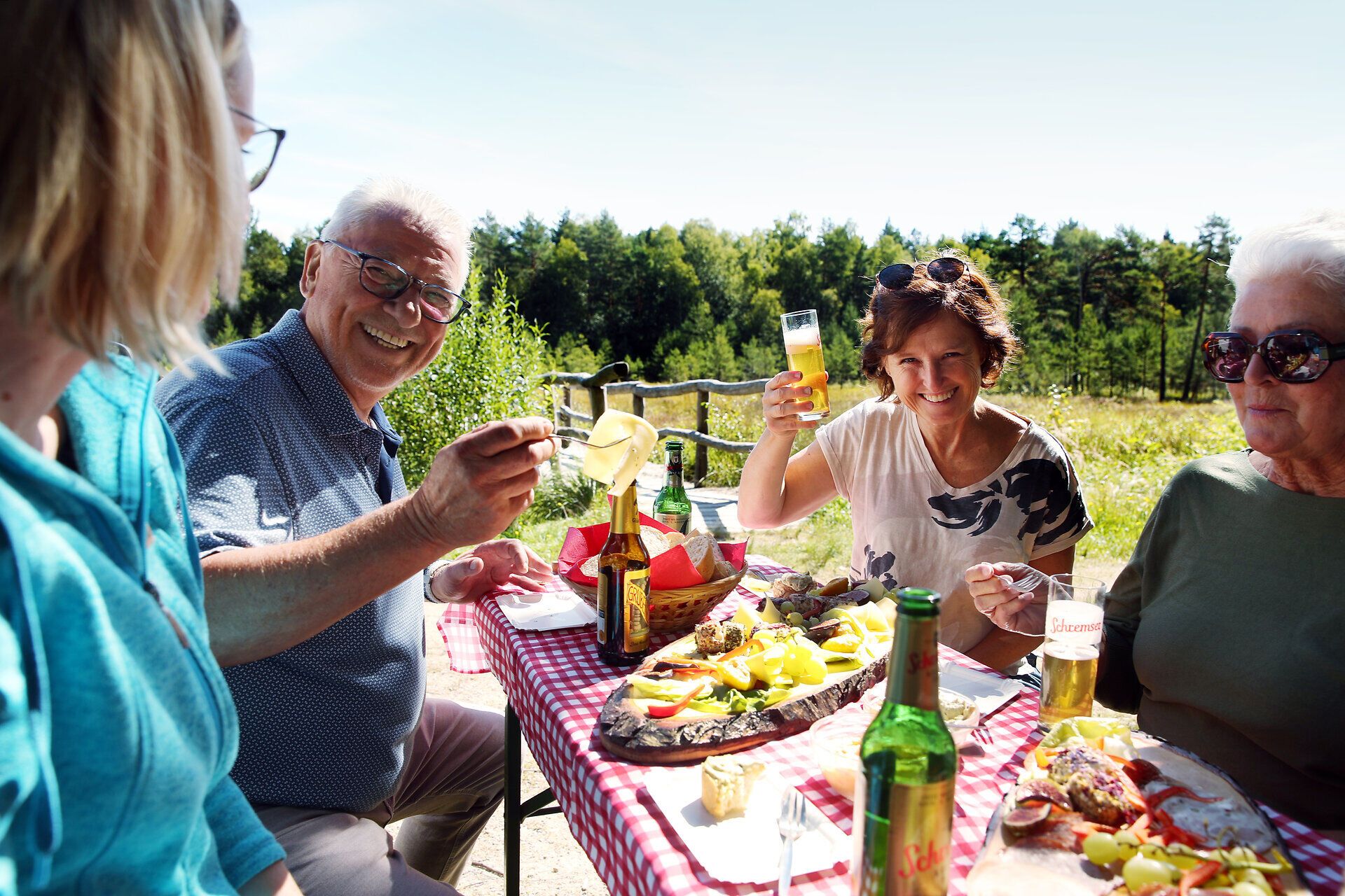Eine Gruppe genießt das Angebot "Moor, Käse und Bier" - ein Mann hat ein Stück Käse aufgespießt während eine Frau mit einem Glas Bier anprostet. 