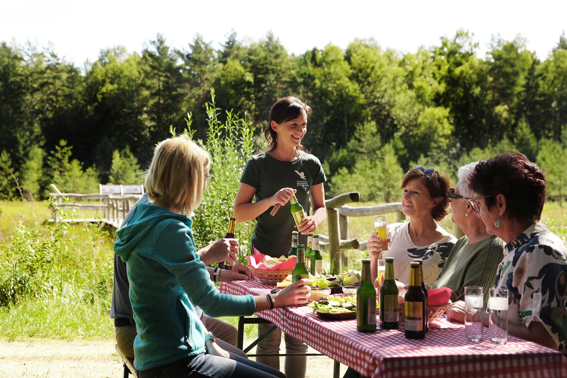 Eine Gruppe genießt das Angebot "Moor, Käse und Bier" und sitzt auf einem Heurigentisch direkt neben dem Prügelsteg.