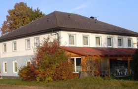 Zweistöckiges Gebäude mit grauem Dach und weißen Wänden, umgeben von Bäumen und Sträuchern.