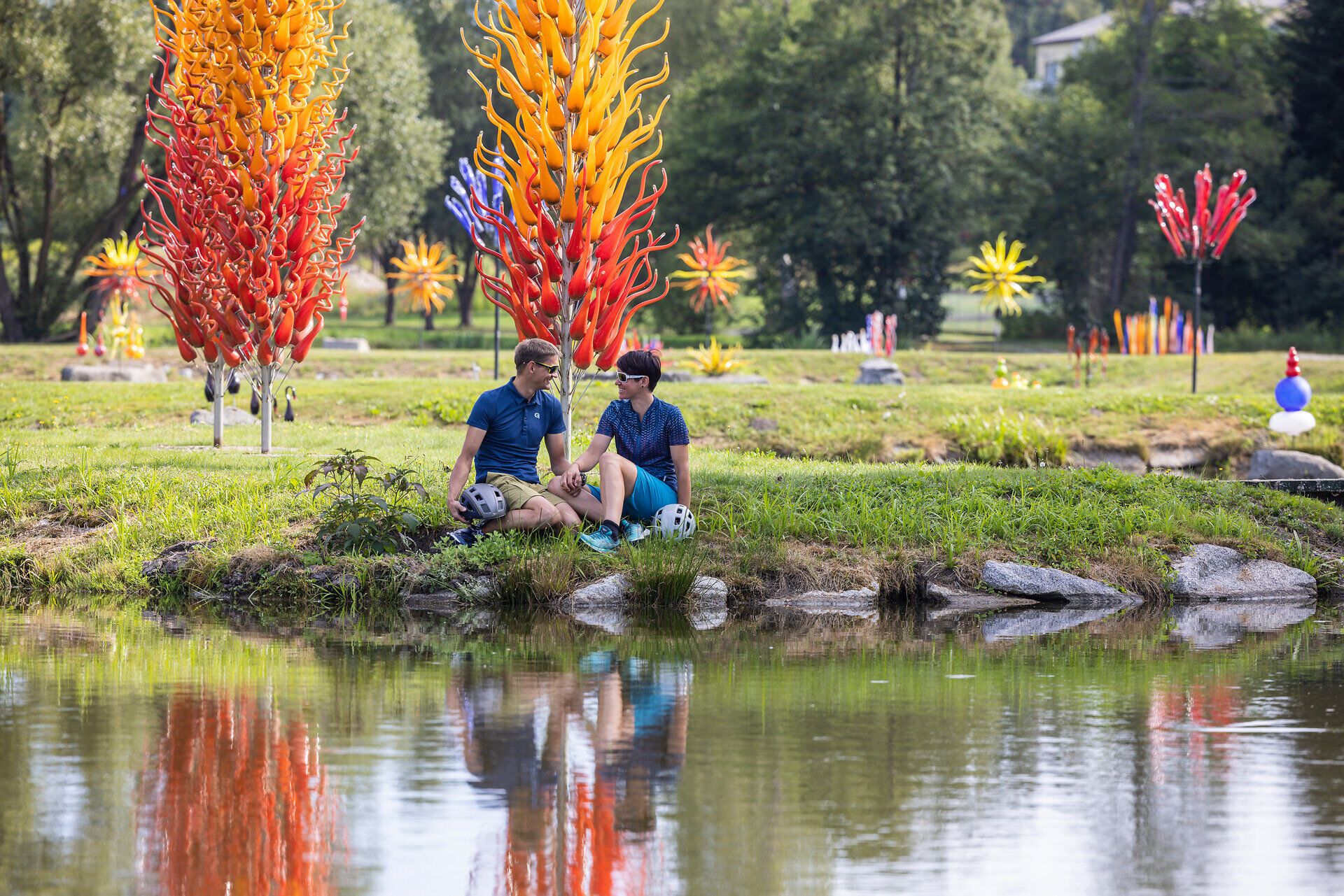 Inmitten einer faszinierenden Wasserlandschaft entspannen zwei Radfahrer am Ufer, umgeben von leuchtenden Glaskunstwerken, die in der Sonne strahlen. Die harmonische Verbindung von Kunst und Natur schafft eine einladende Atmosphäre, die zum Verweilen einlädt und die Schönheit des Waldviertels erlebbar macht.