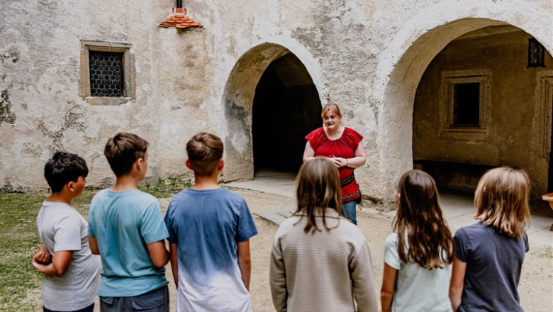 Eine Frau in rotem Oberteil spricht zu einer Gruppe von Kindern vor einer alten Steinmauer mit Torb&ouml;gen.
