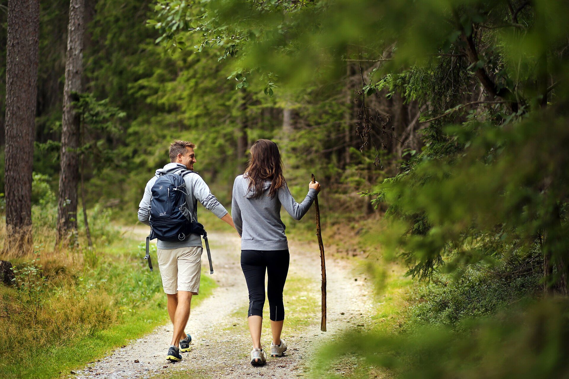 Ein Paar wandert entspannt durch den dichten Wald, umgeben von hohen Bäumen und dem sanften Licht, das durch das Blätterdach fällt. Die frische Luft und das Zwitschern der Vögel schaffen eine harmonische Atmosphäre, die zum Verweilen einlädt.
