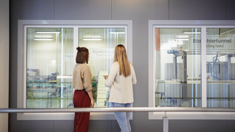 Zwei Frauen betrachten durch ein Fenster eine Produktionsanlage.