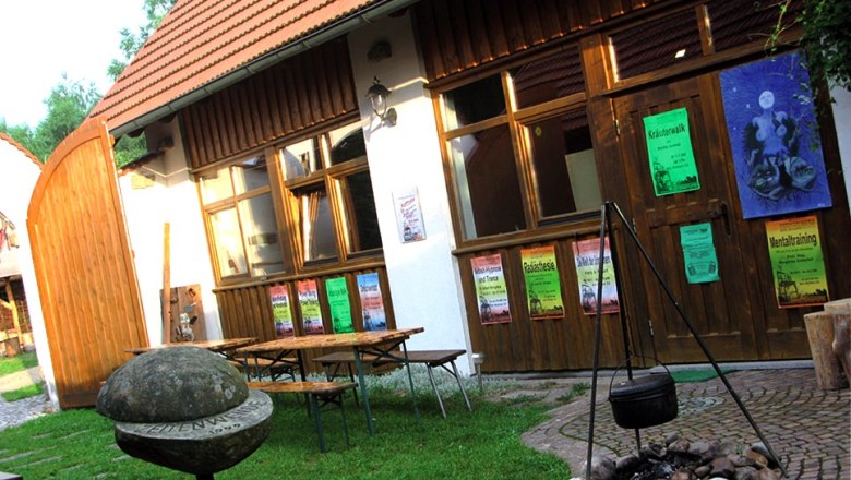 Ein Hof mit Holztischen, Plakaten an einer Holzwand und einem Kessel über einem Feuerplatz.
