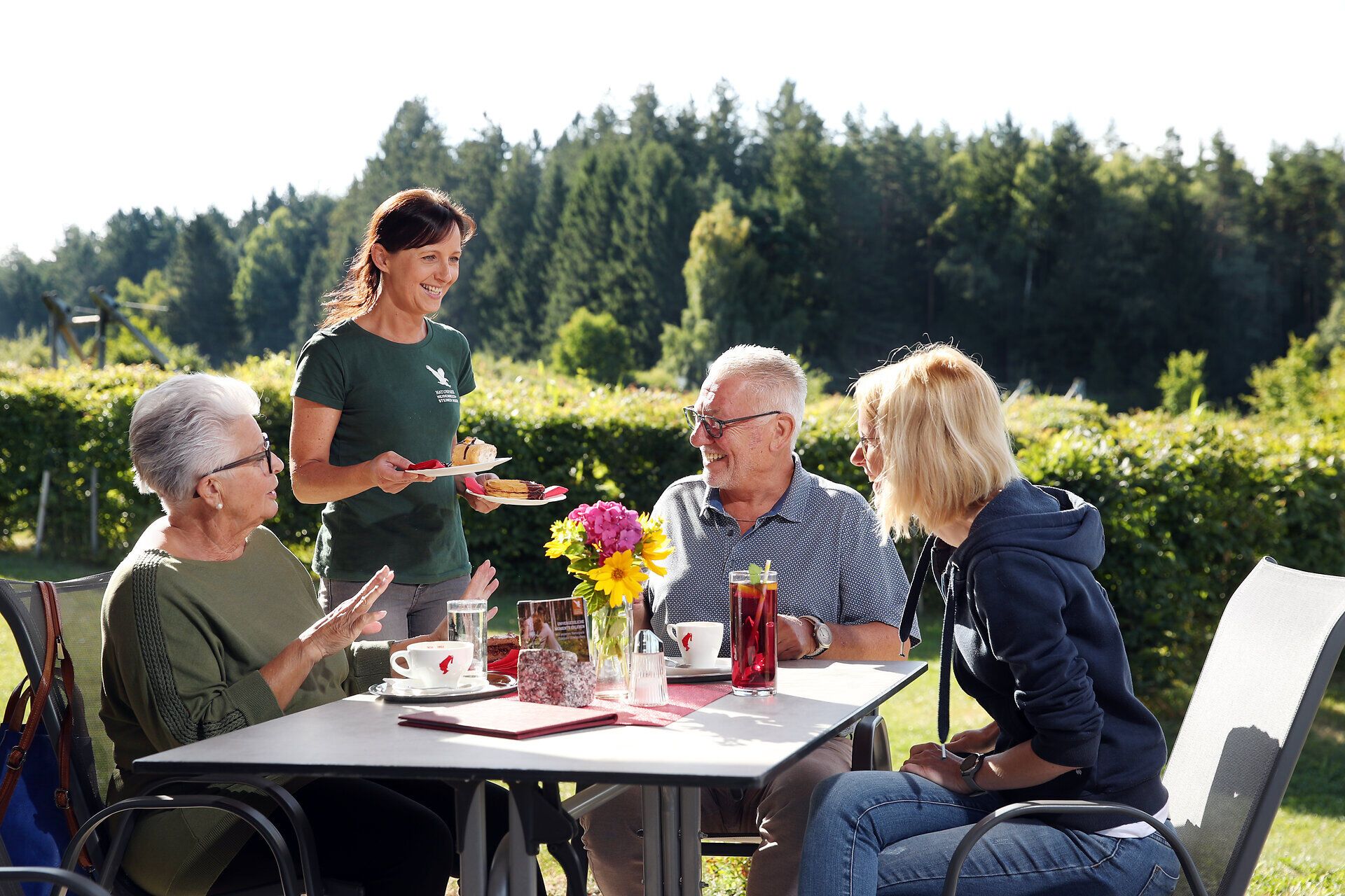 Drei Personen sitzen gemütlich auf der Sonnterasse beim Naturpark-Zentrum in Heidenreichstein beisammen, eine Damen serviert Mehlspeisen. 